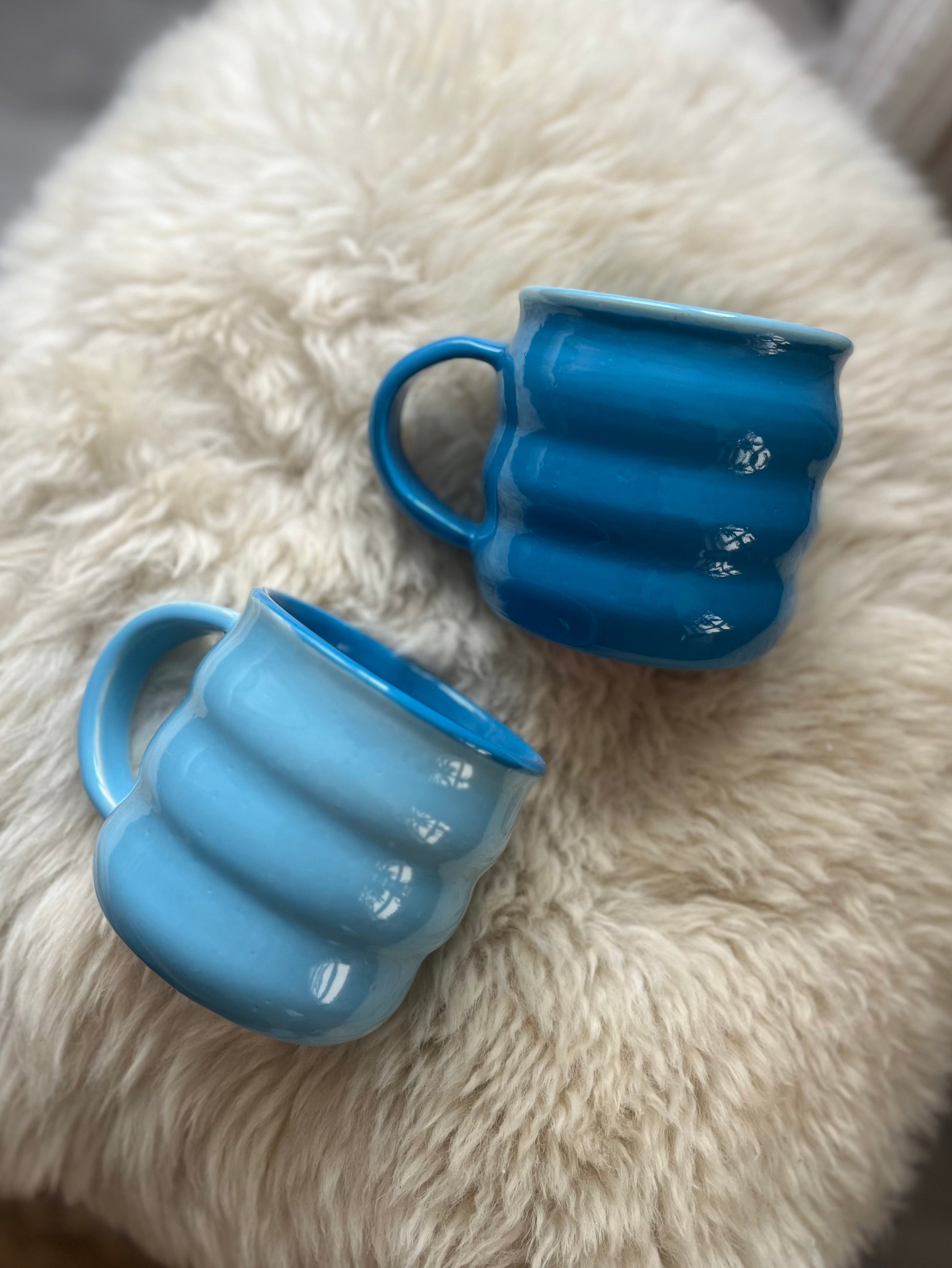 Two ribbed ceramic mugs in light blue and dark blue, displayed on a fluffy white faux fur surface.