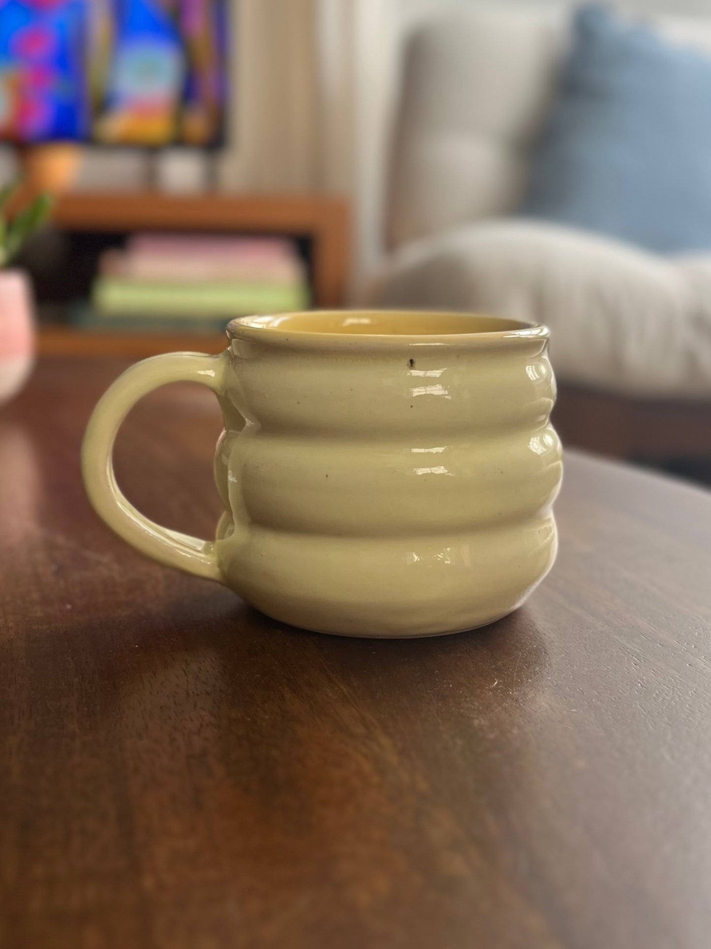 A pale yellow-green ceramic mug with a ribbed, wavy design and a loop handle, sitting on a dark wooden table in a blurred living room setting.