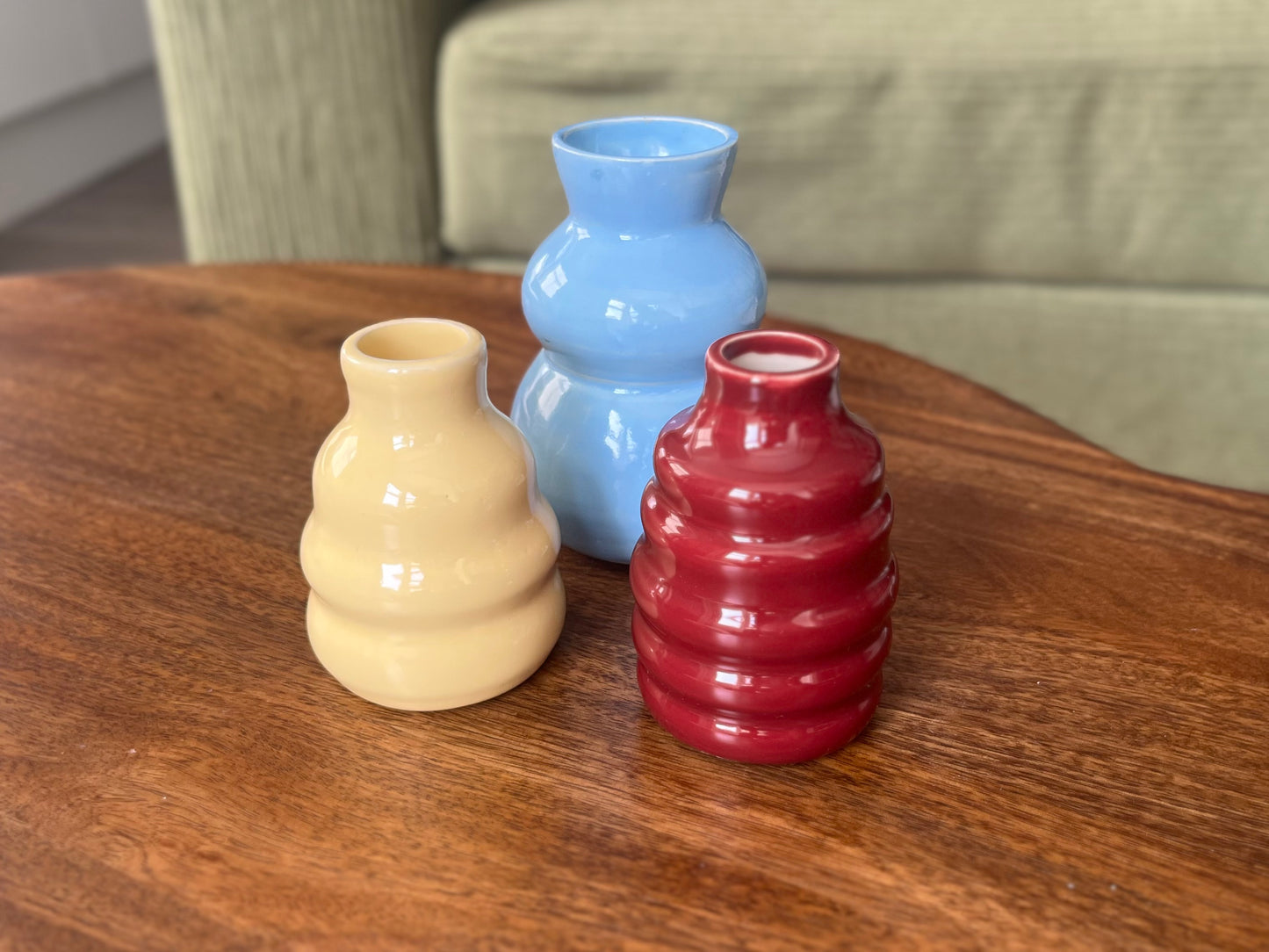 Three small ceramic bud vases in yellow, light blue, and dark red, arranged on a wooden table with a blurred sofa in the background.