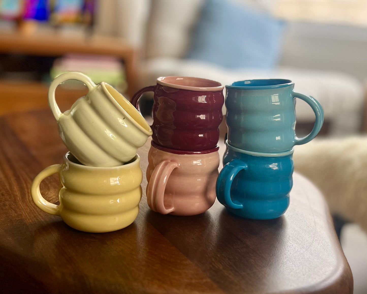Four stacked ceramic mugs in pastel and rich colors, including yellow, maroon, light blue, and pink, arranged on a dark wooden table in a living room setting.