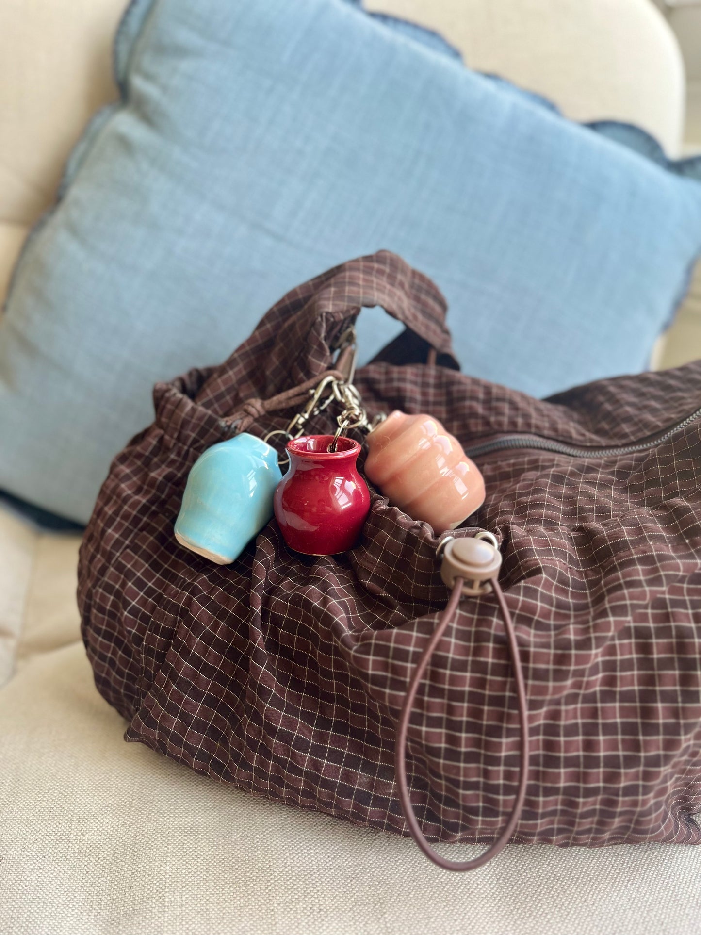 A brown plaid fabric shoulder bag with a zipper closure, resting on a beige sofa with a blue pillow in the background. The bag is decorated with three small ceramic mini vase charms in light blue, burgundy, and pink, attached by a stainless steel clip