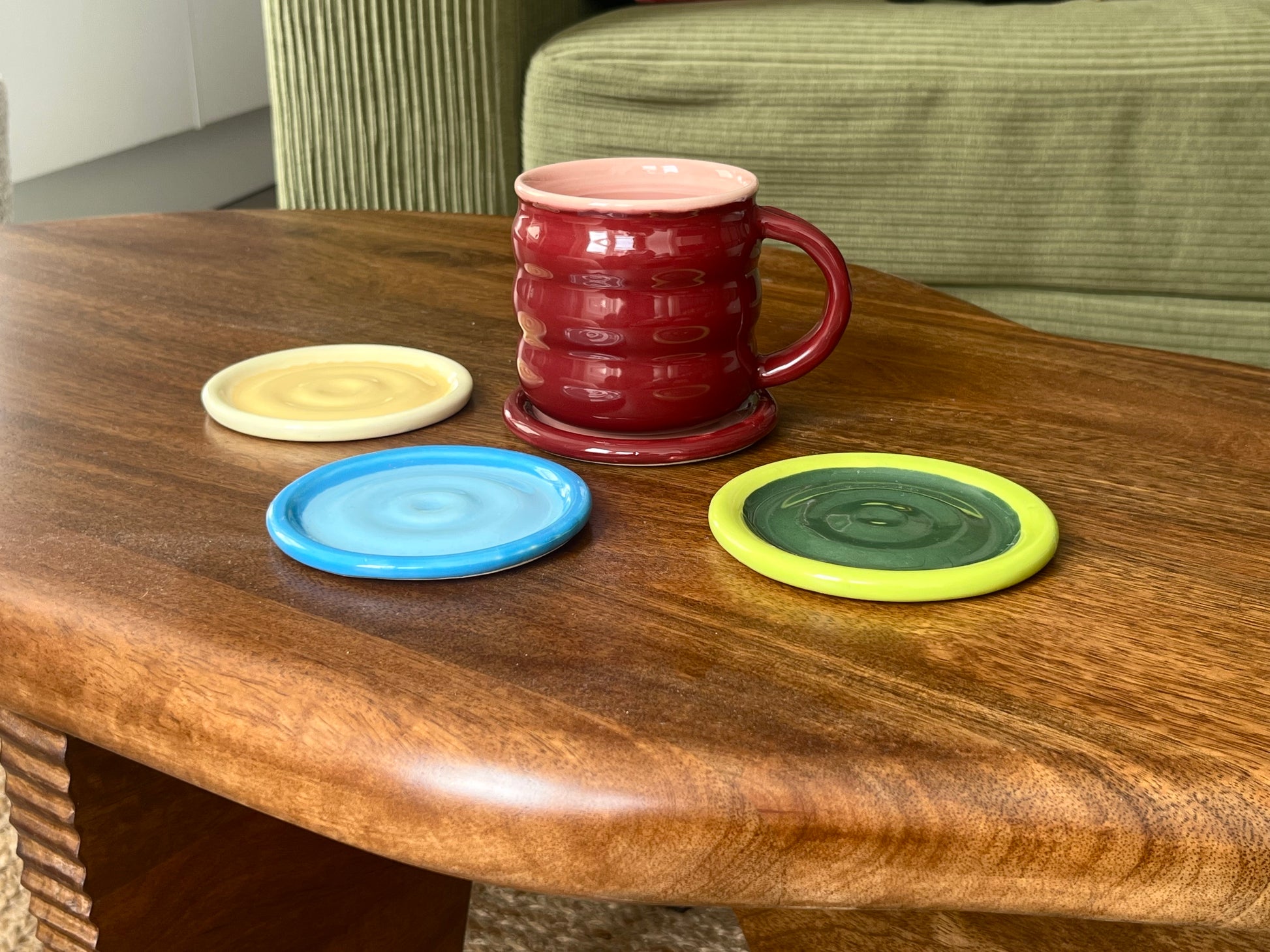 A red ribbed ceramic mug with a pink interior sits on a matching red coaster on a wooden table, alongside three round coasters in yellow, blue, and green.